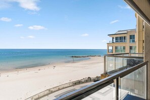 A beach view from a balcony with a clear sky, a sandy beach, and a building with multiple floors and balconies. - Oceanside Studio at the Pier (Holdfast Bay)