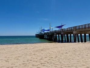 Ein Strand mit einem Steg, der sich ins Meer erstreckt, mit einem Gehweg und Sonnenschirmen.