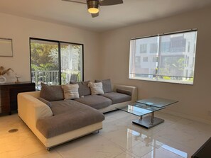 A modern living room with a sofa, a glass coffee table, and large windows offering a view of greenery and buildings. - Bright 2BR Coastal Suite Apt12 (Pompano Beach)
