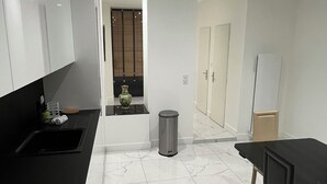 A modern kitchen with white cabinets, a black countertop, a wooden cutting board, a trash can, and a dining area with a table and chairs. - Peaceful and spacious T3 in the heart of Bordeaux (Bordeaux)