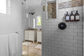 A modern bathroom with a glass shower enclosure, white tiled walls, a wooden vanity, and three bottles of toiletries.
