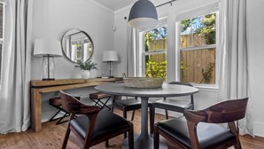 A dining area with a round table, four chairs, a lamp, a mirror, and a window with a view of greenery.