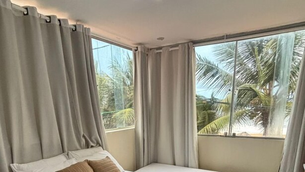 A bedroom with two beds, a large window with curtains, a ceiling light, and a view of palm trees. - Pousada Recanto Paradise  (Ipojuca)