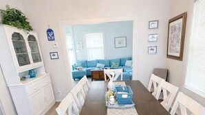 Ein Essbereich mit einem langen Tisch, weißen Stühlen, einer Chandelierleuchtre und Blick in ein Wohnzimmer mit einem blauen Sofa.