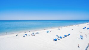 A beach with white sand, clear blue water, and several blue umbrellas. - 603 Villa Madeira White Sand Beachfront Condo with Stunning Sunsets (Madeira Beach)