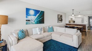 A living room with a sectional sofa, a blue rug, a painting of a wave, and a dining area with a table and chairs. - 603 Villa Madeira White Sand Beachfront Condo with Stunning Sunsets (Madeira Beach)