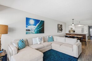 Ein Wohnzimmer mit einem Ecksofa, einem blauen Teppich, einem Gemälde einer Welle und einem Essbereich mit Tisch und Stühlen.
