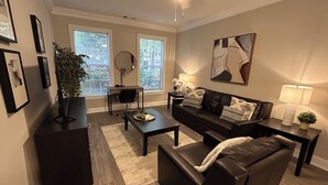 A living room with a sofa, coffee table, and a desk with a chair. There are windows, a plant, and artwork on the walls. - Spacious Corporate Furnished One Bedroom/One Bath in Alpharetta. (Alpharetta)