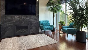 Ein modernes Wohnzimmer mit Flachbildfernseher an einer dunklen Marmorwand, Kamin, Holzboden, petrolfarbenen Sesseln und großen Fenstern.