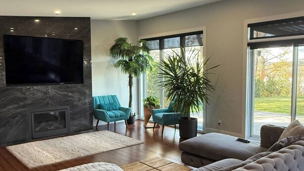 Ein modernes Wohnzimmer mit einem großen Flachbildfernseher, einem Kamin, einem Ecksofa und einem großen Fenster mit Blick ins Freie.