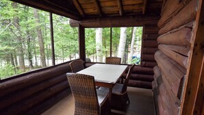 A log cabin with a dining table and wicker chairs, large windows, and a view of a forest. - Cozy Adirondack Cabin-Lakeside Resort Camp 1 (Olmstedville)