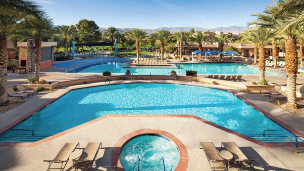 Una gran piscina exterior con varias secciones, rodeada de palmeras y sillones de descanso.