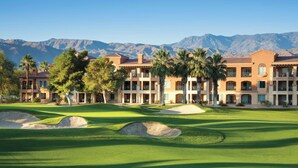 Un campo de golf con búnkeres y palmeras, un edificio con balcones, y montañas al fondo.
