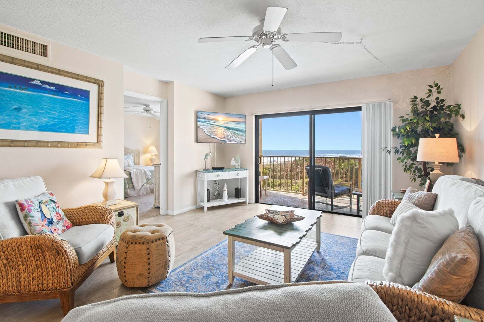 A spacious living room with a ceiling fan, a large window with a view of the ocean, a dining area with a table and chairs, and a balcony with a chair.