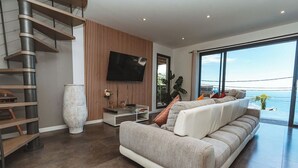 Een moderne woonkamer met een grote bank, een aan de muur bevestigde televisie en uitzicht op zee door grote ramen.