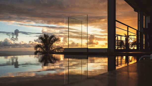 Een modern gebouw met een glazen gevel die een zonsondergang over het water, palmbomen en een brug weerspiegelt.