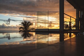 En moderne bygning med en glasfacade, der spejler en solnedgang over vandet, palmer og en bro.