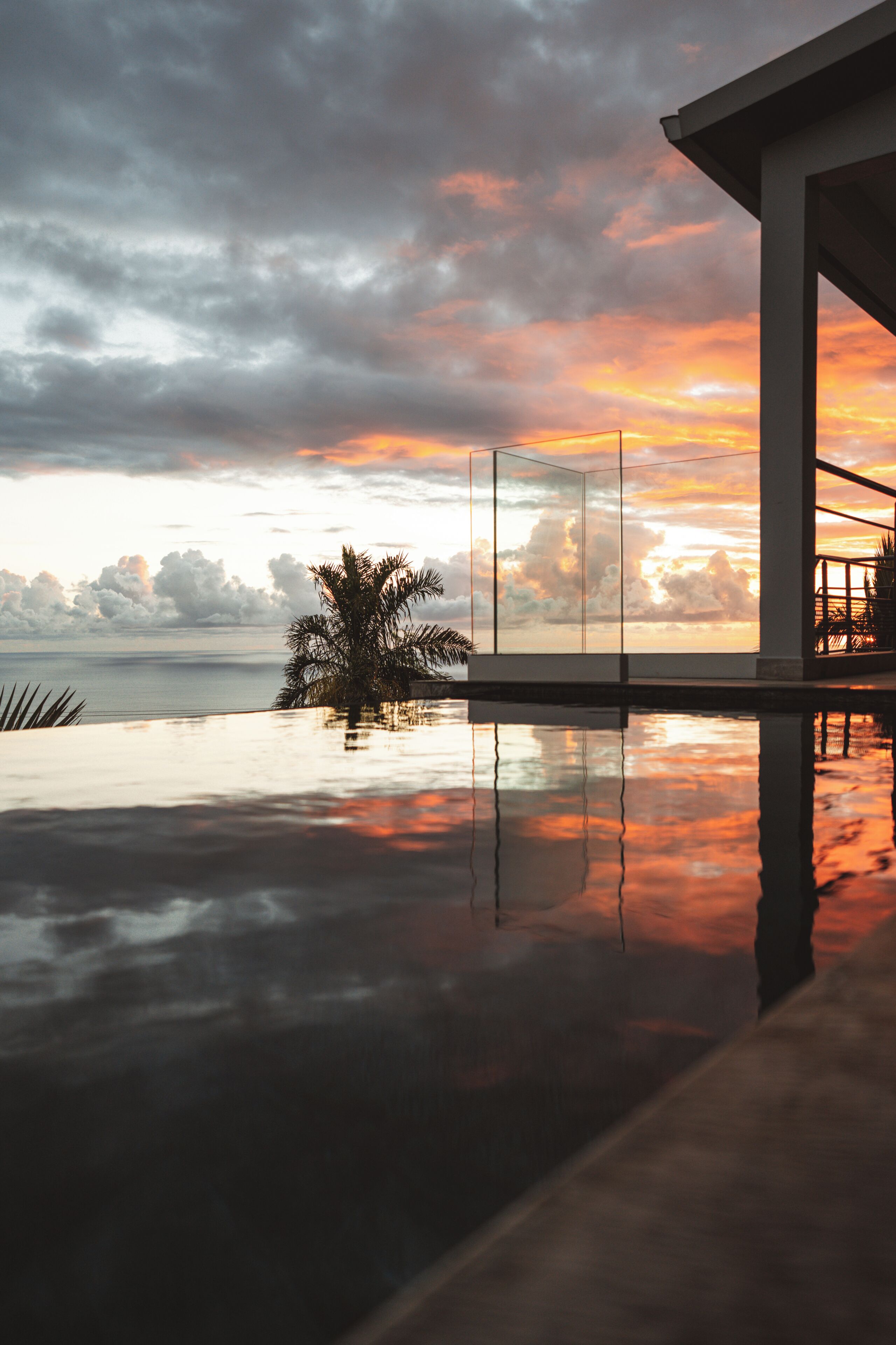 Une piscine à débordement moderne qui reflète le ciel au coucher du soleil, un palmier et une structure architecturale.