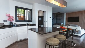 Een moderne keuken met een centraal eiland, barkrukken, een eettafel met fruit en een aan de muur bevestigde televisie.
