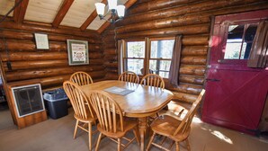 A wooden cabin interior with a dining table, chairs, a red door, and a window with curtains. - Cozy Adirondack Cabin-Lakeside Resort Camp 3 (Olmstedville)