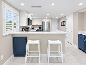 A modern kitchen with white cabinets, a stainless steel refrigerator, and a built-in microwave. There are two white bar stools and a dark blue cabinet. - Beachfront Condo Sanibel Sandalfoot 1A2 (Sanibel Island)