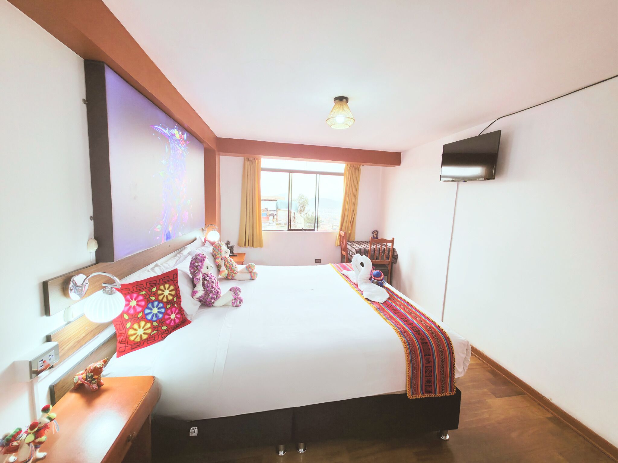 Habitación de hotel con una cama grande, una mesita de noche con lámpara, un televisor montado en la pared y una ventana con cortinas.