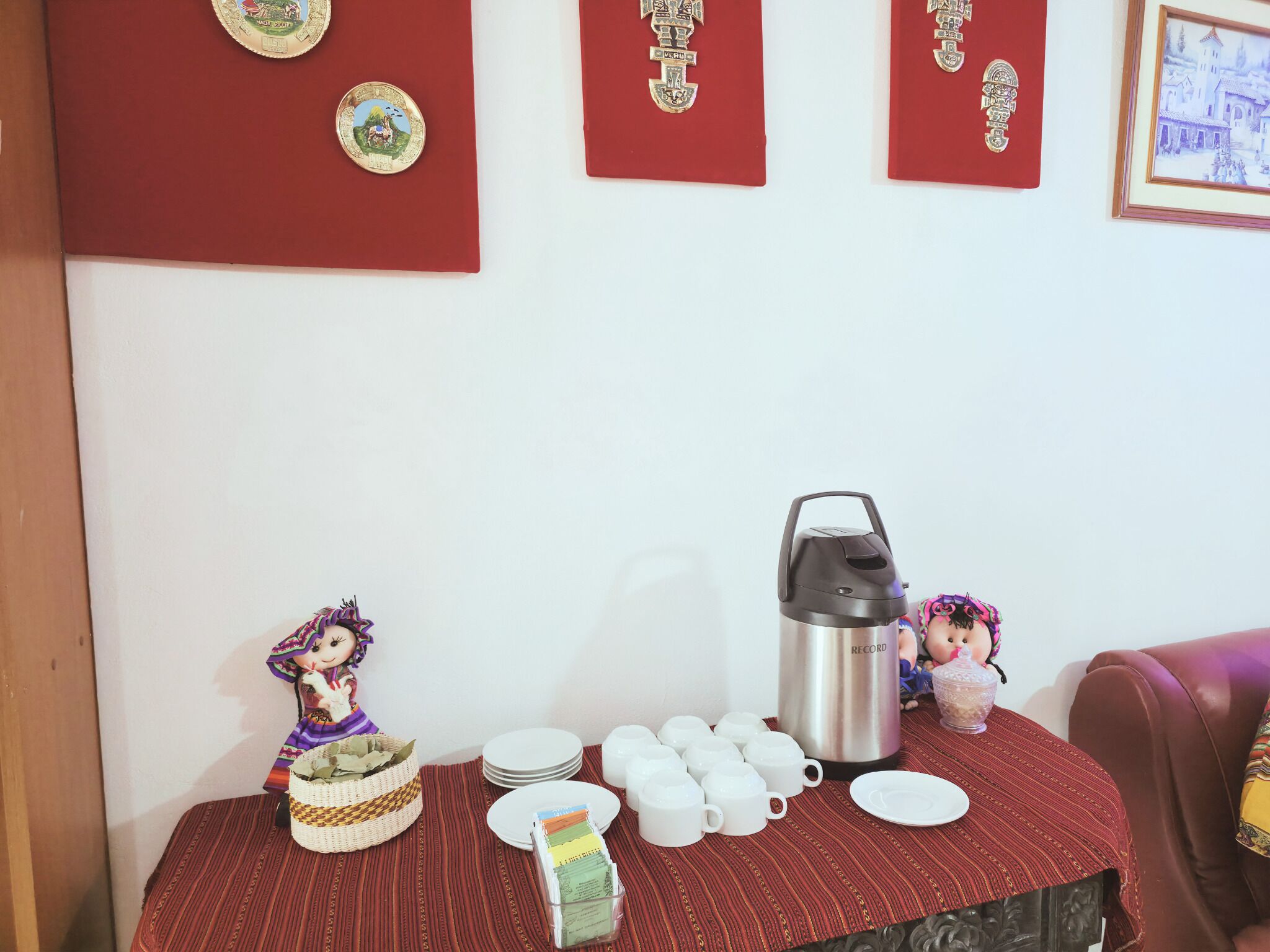 Une table recouverte d’une nappe rouge, sur laquelle on trouve une machine à café, des tasses, des assiettes, une poupée et divers objets de décoration.