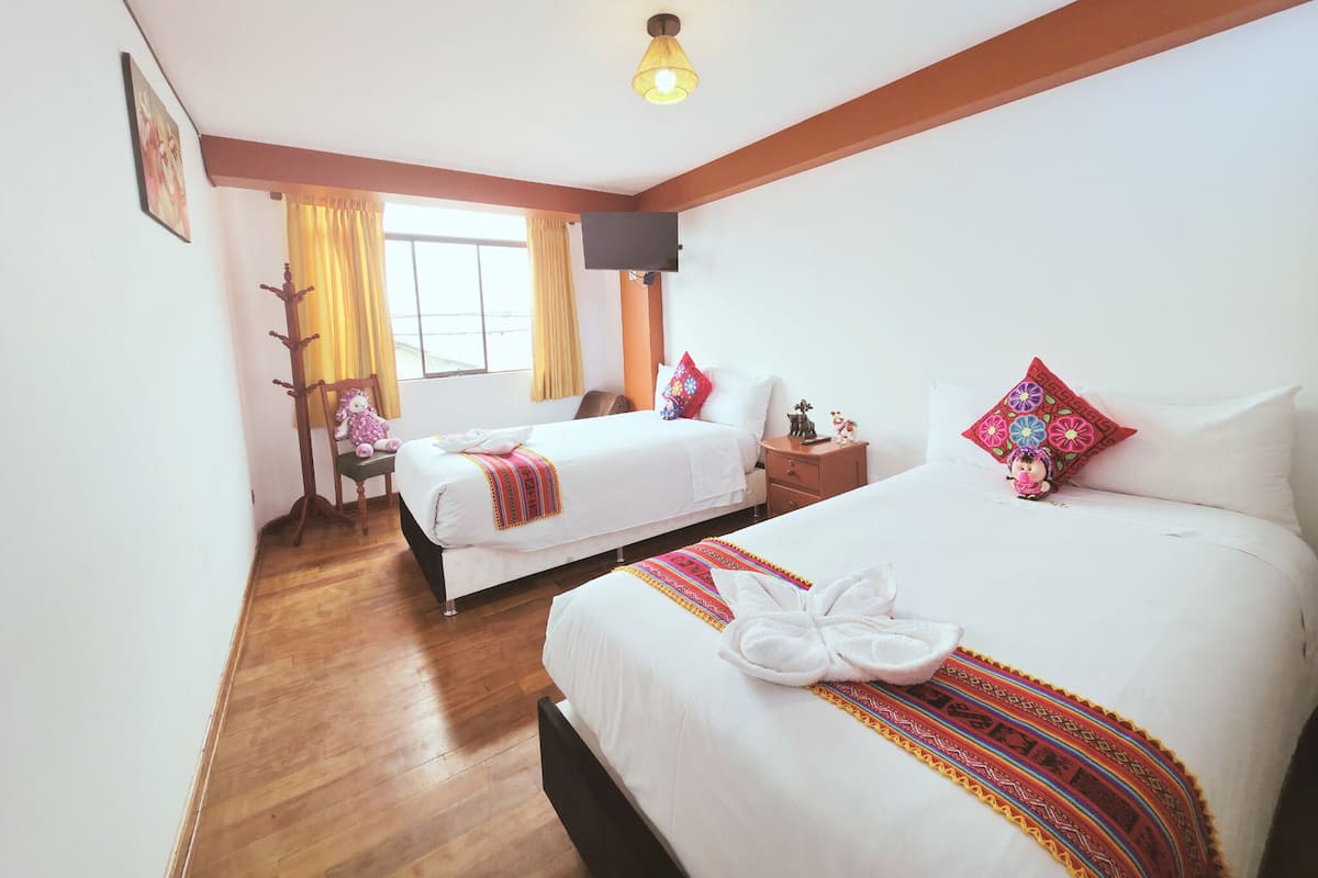 A hotel room with two beds, a wooden floor, a window with curtains, a flat-screen TV, and a wall-mounted light fixture.