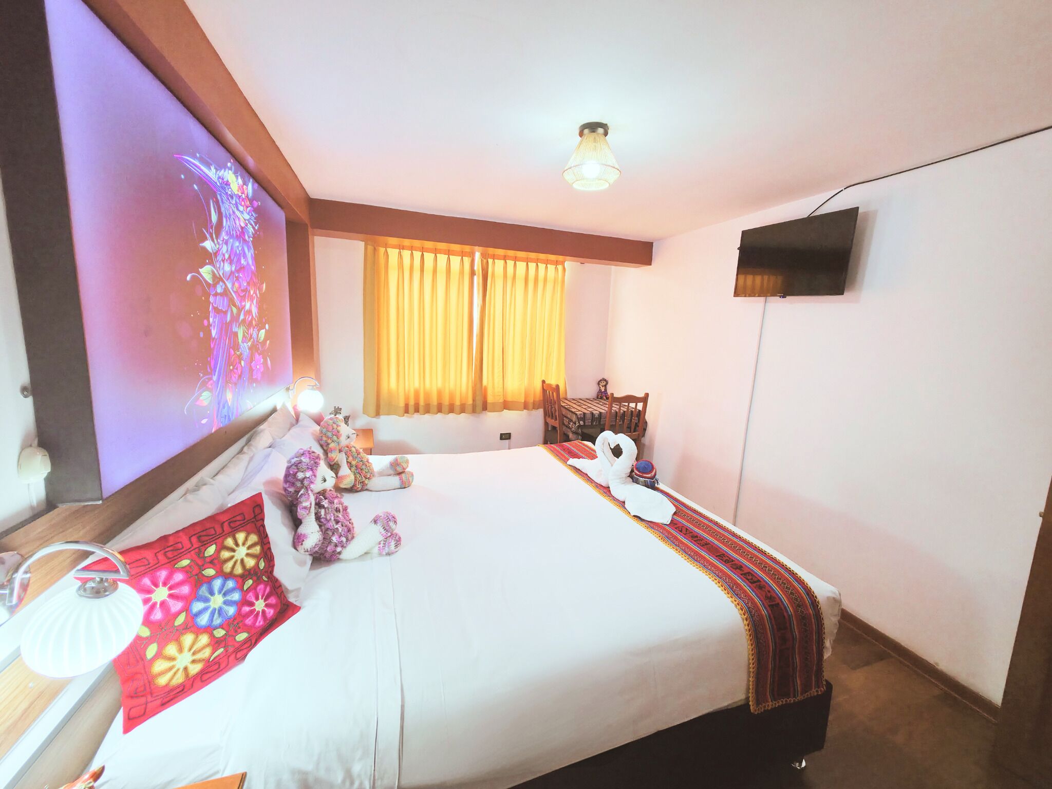 Habitación de hotel con cama, televisión montada en la pared, ventana con cortinas y mesita de noche con lámpara.