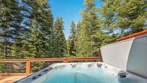 Een hottub met een houten terras en een metalen reling, omgeven door hoge bomen.