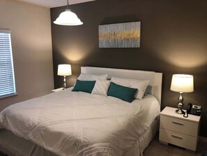 A bedroom with a bed, bedside lamps, a nightstand, a wall-mounted artwork, and a window with blinds. - Comfortable 3BR Townhome Near Parks, Great Value (Kissimmee)