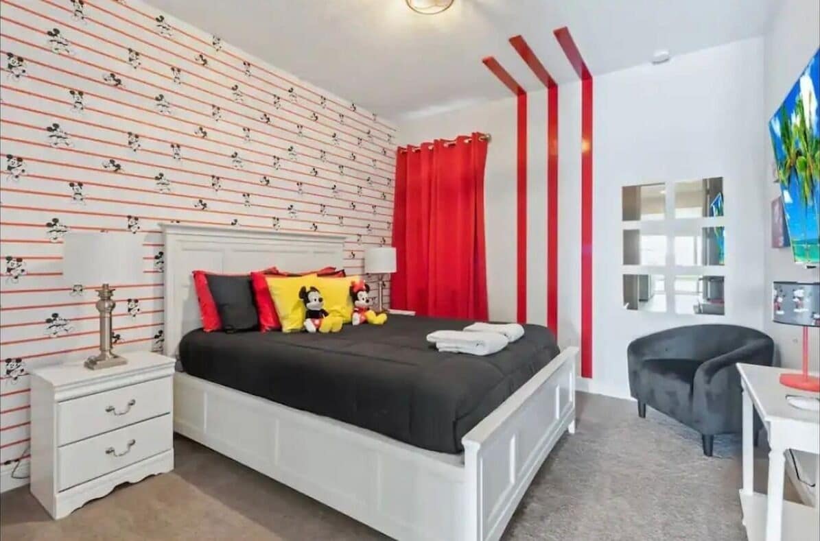Un dormitorio con una cama, una mesita de noche, una silla y una pared con estampado de Mickey Mouse.