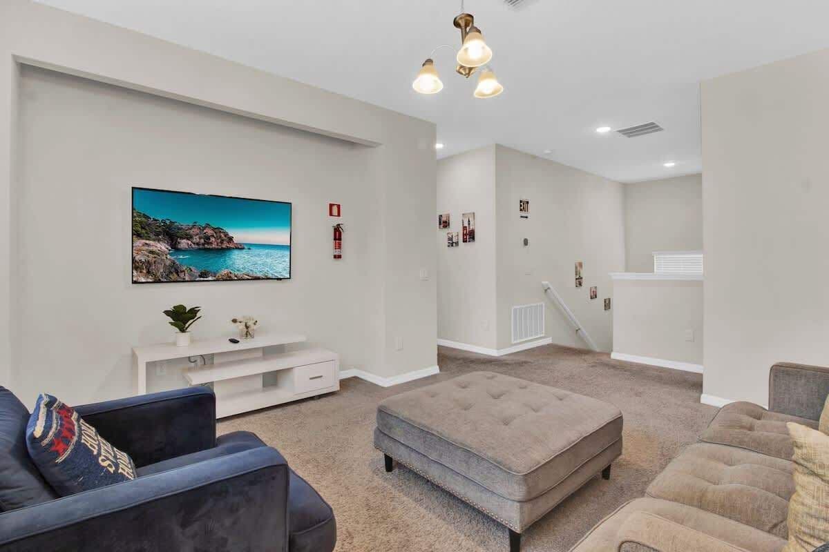 A living room with a large flat-screen TV, a tufted ottoman, a blue sofa, and a beige sofa.