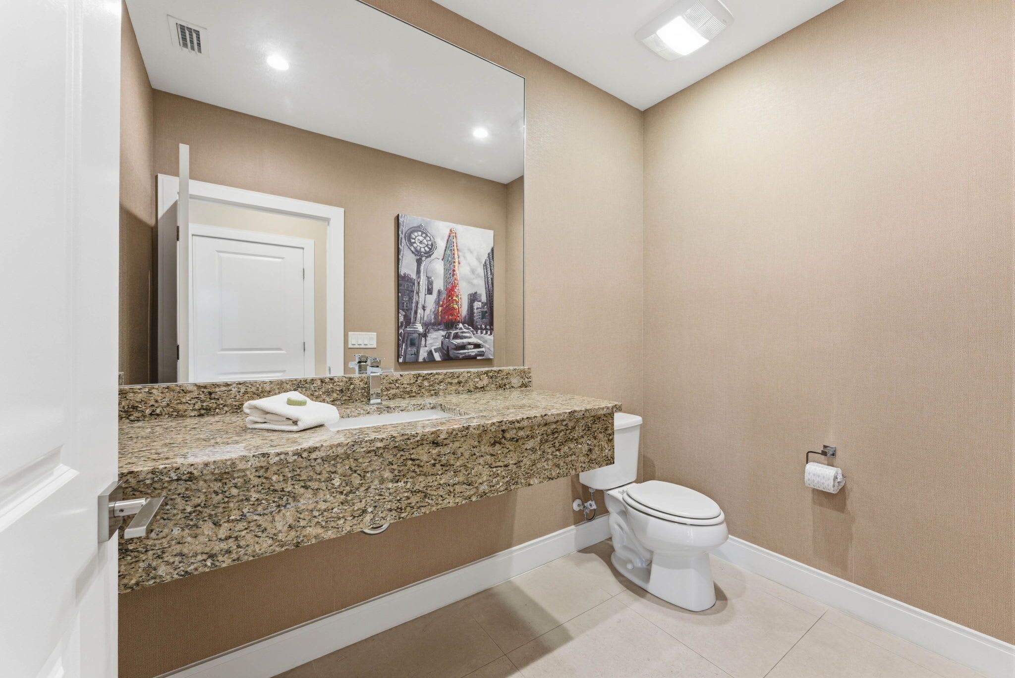 Une salle de bain avec un comptoir en granit, des toilettes blanches, un grand miroir et un tableau accroché au mur.