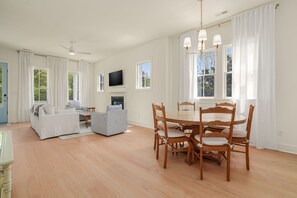 A spacious living room with a sofa, armchair, and a dining table with chairs. A flat-screen TV is mounted on the wall, and there is a fireplace.