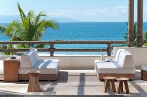 A balcony with a sofa, two wooden stools, and a small table, overlooking a beach with palm trees and the ocean. - LUXE BEACHFRONT FULLY STAFFED WITH CHEF AT VENEROS (Bahia de Banderas)