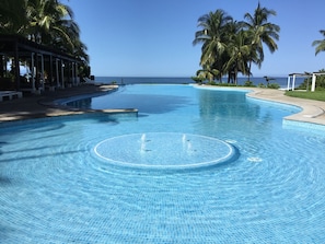 Ein klares, blaues Infinity-Pool mit einem runden Wasserspiel, umgeben von Palmen und Blick auf das Meer.