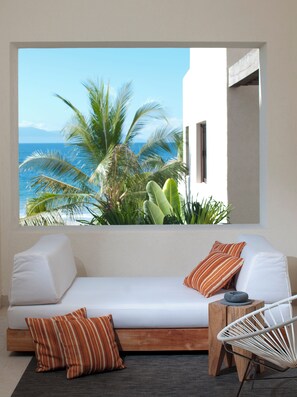Ein Zimmer mit einem weißen Sofa, gestreiftem Kissen, einem Korbstuhl und Blick auf das Meer und Palmen.