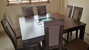 Uma mesa retangular para refeições com seis cadeiras, uma janela com grades e uma parede lisa.