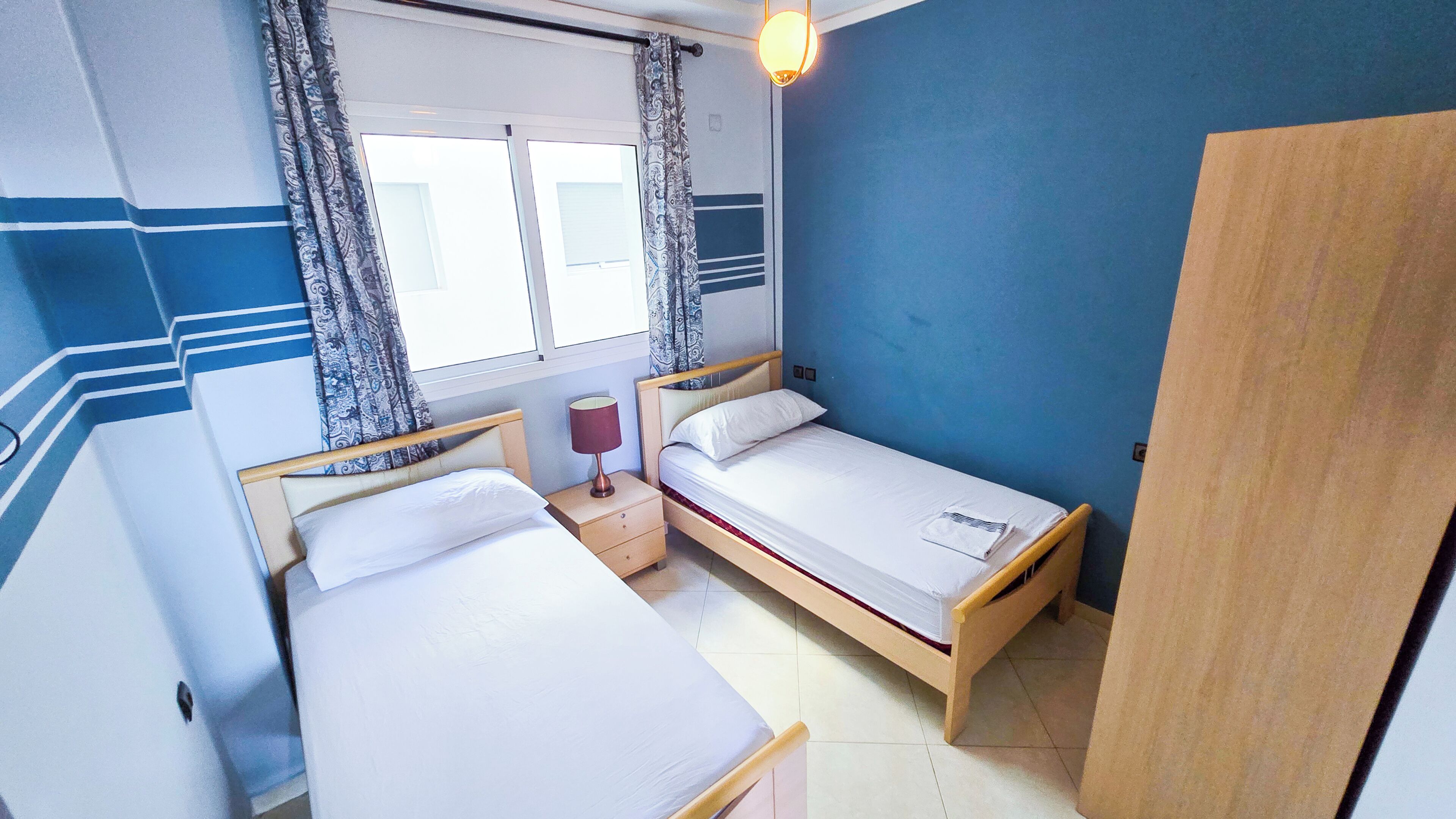 Una habitación con dos camas individuales, una mesita de noche con lámpara, un armario y una ventana con cortinas.