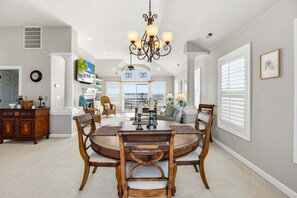 A spacious dining area with a round table, wicker chairs, and a chandelier. - Bay Dreams Soundfront Escape w/ Beach Parking & Sunsets (Kill Devil Hills)