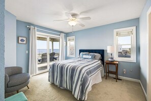A bedroom with a bed, a chair, a nightstand, a ceiling fan, and a window. - Bay Dreams Soundfront Escape w/ Beach Parking & Sunsets (Kill Devil Hills)
