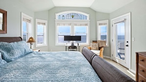 Ein Schlafzimmer mit einem großen Bett, einem Schreibtisch mit Computer, einem Sessel und Meerblick durch ein Fenster.