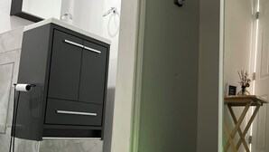 Une salle de bain moderne avec des toilettes, un lavabo et un miroir. On y trouve un porte-serviettes et une petite table en bois sur laquelle est posé un vase. Le sol est carrelé et les murs sont peints en blanc.