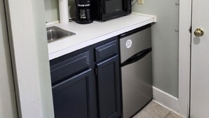 A compact kitchen with white cabinets, a black microwave, a coffee maker, and a stainless steel refrigerator.