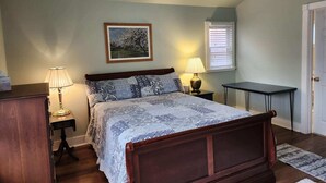 A bedroom with a bed, two bedside tables with lamps, a framed picture on the wall, a window with blinds, and a door.