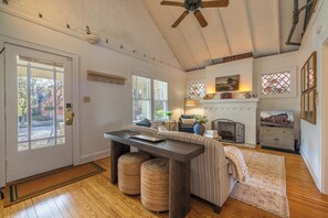 A room with a wooden floor, a ceiling fan, a fireplace, and a window with a view of trees.