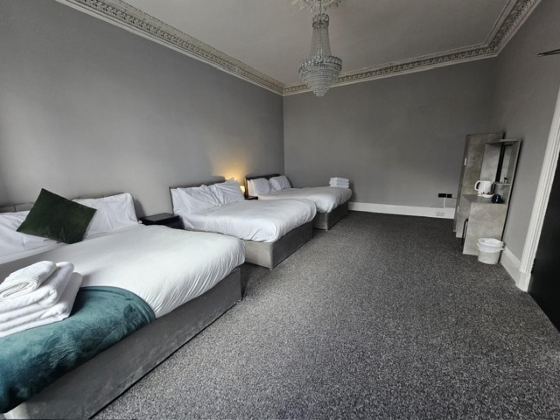 A hotel room with three beds, a chandelier, a mirror, and a small bathroom area.