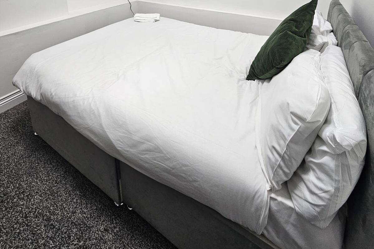 A neatly made bed with white bedding, a grey headboard, and a single green pillow. A flat-screen TV is mounted on the wall above the bed.