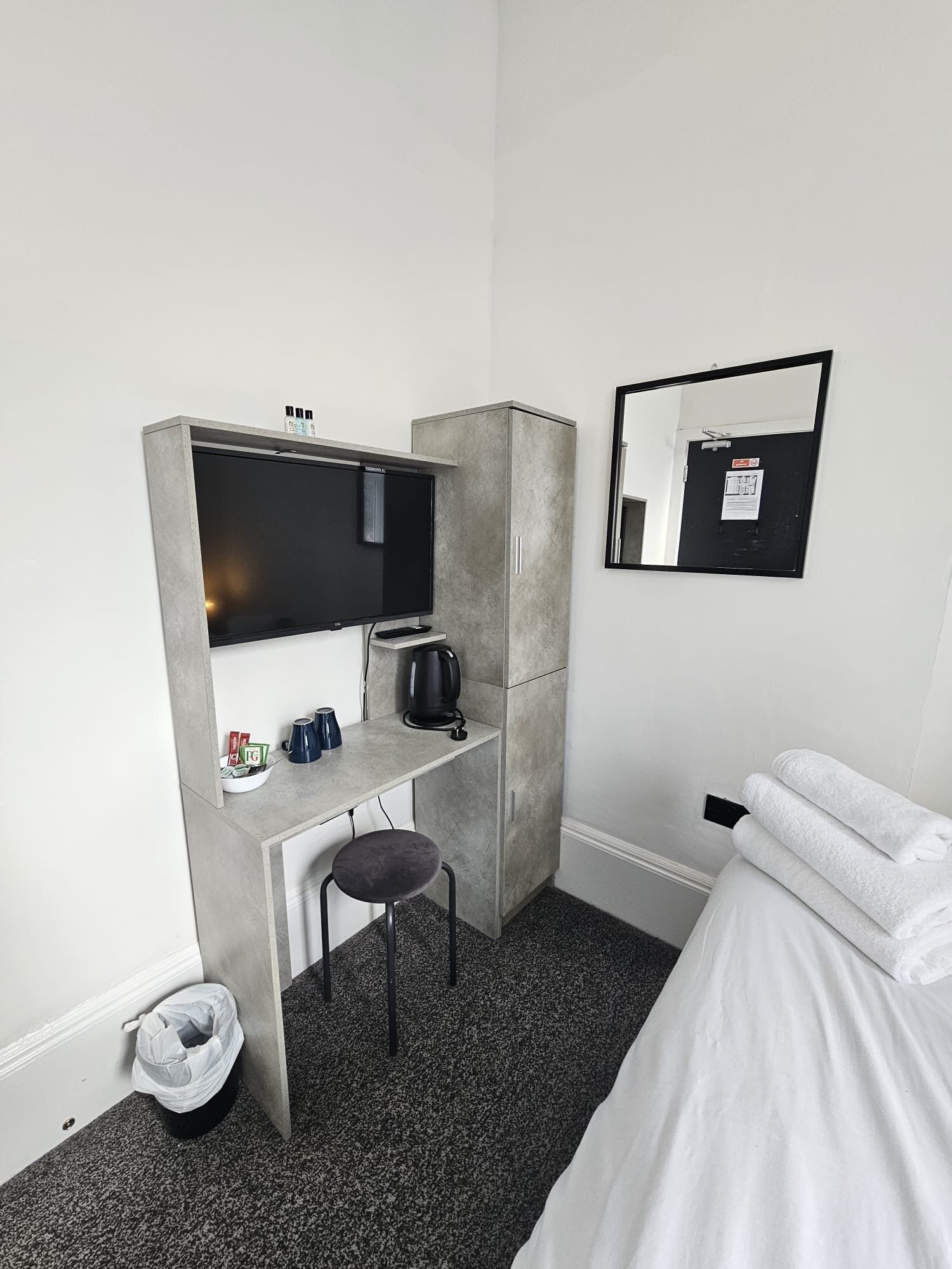 Quarto de hotel compacto com cama, escrivaninha com televisão, um banquinho, lixeira e espelho.